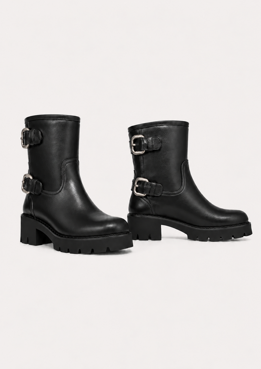 A pair of black leather ankle boots with chunky lug soles and buckle straps on the side, displayed against a light gray studio background.