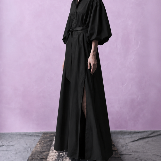 A model wearing a long black shirt dress with voluminous sleeves and a waist tie, standing on a rustic wooden platform against a textured lavender-purple studio backdrop.