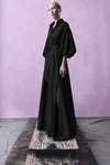 A model wearing a long black shirt dress with voluminous sleeves and a waist tie, standing on a rustic wooden platform against a textured lavender-purple studio backdrop.