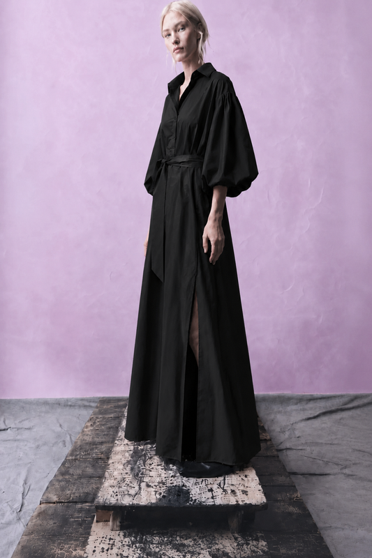 A model wearing a long black shirt dress with voluminous sleeves and a waist tie, standing on a rustic wooden platform against a textured lavender-purple studio backdrop.