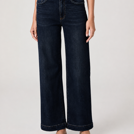 A model wearing dark blue wide-leg denim jeans with decorative chain stitching along the front pockets, paired with a white top and white slide sandals, against a light gray studio background.