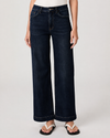 A model wearing dark blue wide-leg denim jeans with decorative chain stitching along the front pockets, paired with a white top and white slide sandals, against a light gray studio background.