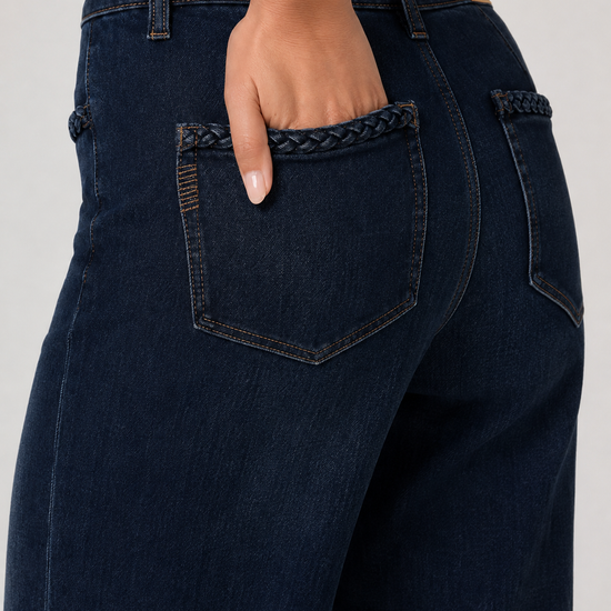 Close-up back view of a person wearing dark indigo jeans with braided pocket trim and a white ribbed top, shown against a light gray studio background.
