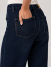 Close-up back view of a person wearing dark indigo jeans with braided pocket trim and a white ribbed top, shown against a light gray studio background.