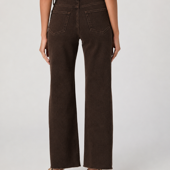Rear view of a person wearing dark brown wide-leg jeans with a white top and tan flats, standing against a light gray studio background.