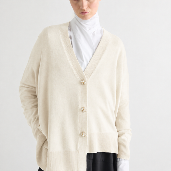 A model wearing a cream-colored knit cardigan with decorative buttons, layered over a white mock neck top and a black skirt, shot against a plain light gray background.