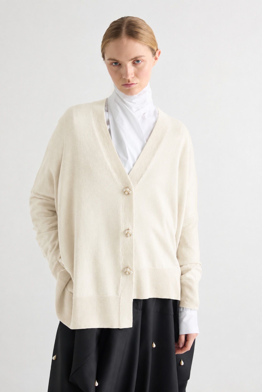 A model wearing a cream-colored knit cardigan with decorative buttons, layered over a white mock neck top and a black skirt, shot against a plain light gray background.