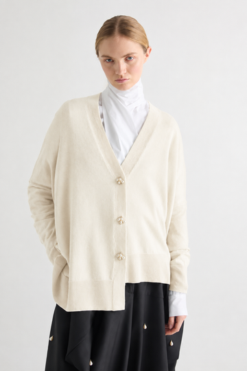 A model wearing a cream-colored knit cardigan with decorative buttons, layered over a white mock neck top and a black skirt, shot against a plain light gray background.