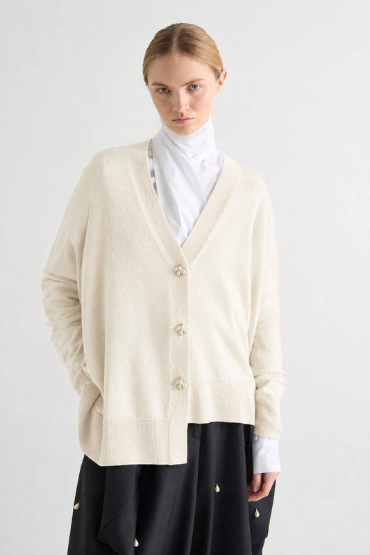 A model wearing a cream-colored knit cardigan with decorative buttons, layered over a white mock neck top and a black skirt, shot against a plain light gray background.