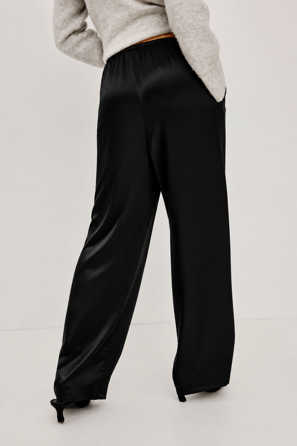 A person wearing black wide-leg satin trousers and a light gray sweater, shown from the back against a plain light gray studio background.