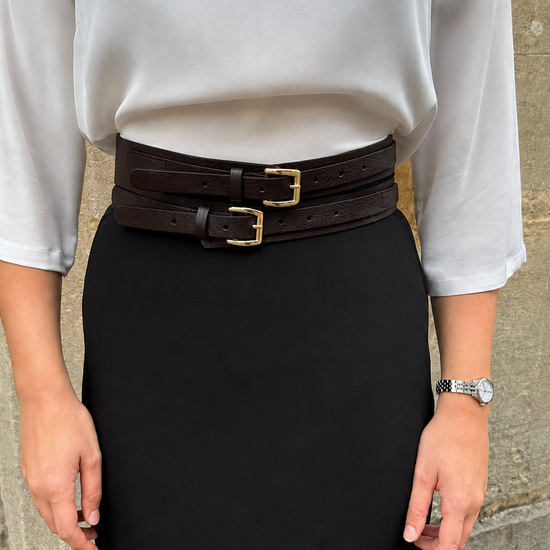 A person wearing a white blouse and a black pencil skirt, styled with a wide dark brown double belt featuring gold-tone buckles, standing against a stone wall.