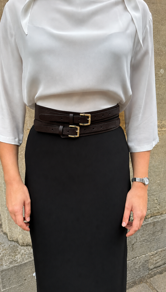 A person wearing a white blouse and a black pencil skirt, styled with a wide dark brown double belt featuring gold-tone buckles, standing against a stone wall.