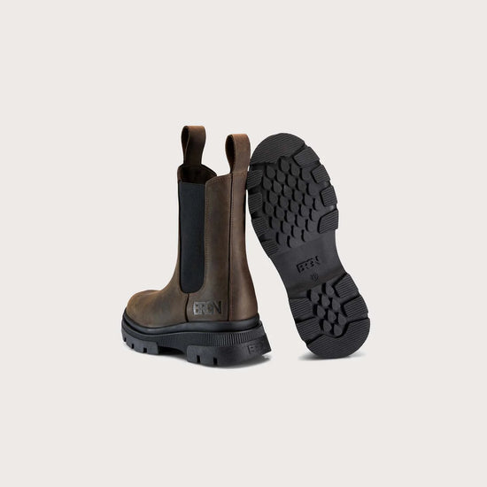 A pair of brown leather Chelsea-style boots shown from the back, with one boot standing upright to display the black elastic side panel and pull tab, and the other lying on its side to show the black lug sole. The brand name 'BRUN' is embossed on the heel and printed on the outsole.