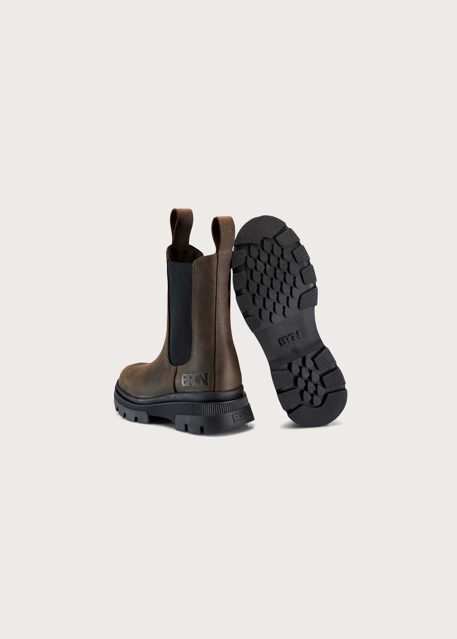 A pair of brown leather Chelsea-style boots shown from the back, with one boot standing upright to display the black elastic side panel and pull tab, and the other lying on its side to show the black lug sole. The brand name 'BRUN' is embossed on the heel and printed on the outsole.