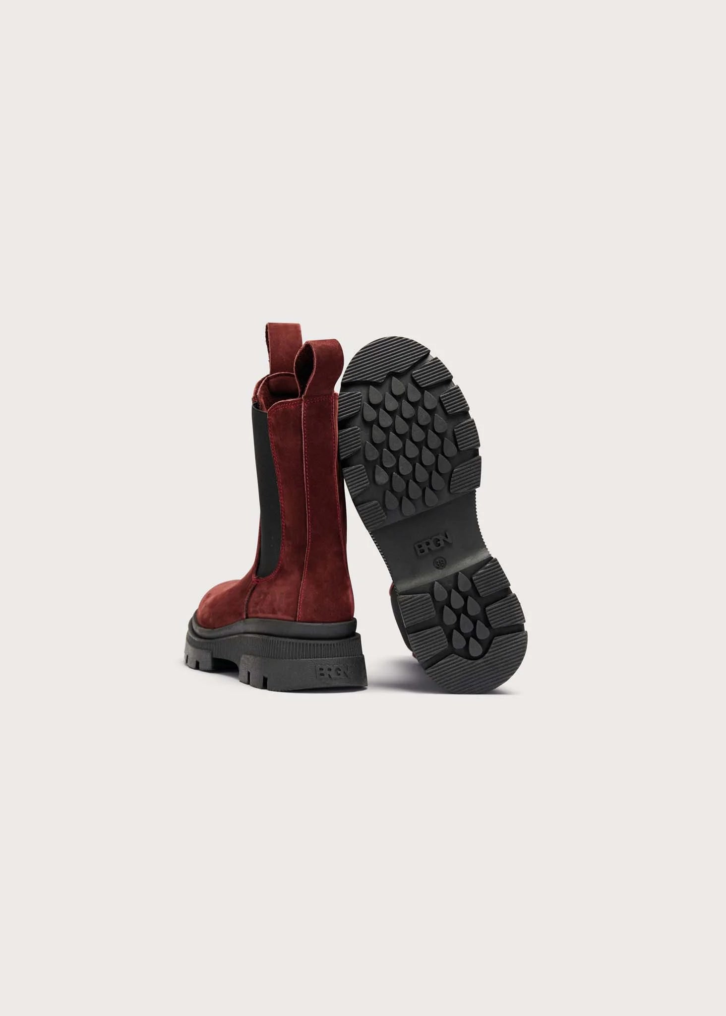 A pair of red suede Chelsea-style boots with black elastic side panels and black lug soles, shown from the back on a light gray background.