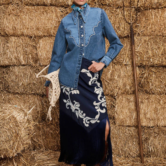Woman in a blue denim shirt and dark blue skirt with white embroidery standing in front of hay bales.