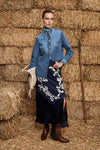 Woman in a blue denim shirt and dark blue skirt with white embroidery standing in front of hay bales.