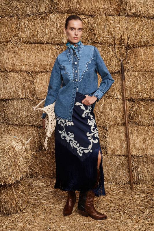Woman in a blue denim shirt and dark blue skirt with white embroidery standing in front of hay bales.