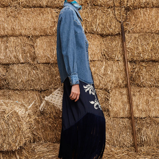 A woman stands in profile against a backdrop of stacked hay bales, wearing a long-sleeve blue denim shirt dress with a dark navy skirt featuring white floral embroidery, paired with brown leather boots.