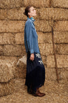 A woman stands in profile against a backdrop of stacked hay bales, wearing a long-sleeve blue denim shirt dress with a dark navy skirt featuring white floral embroidery, paired with brown leather boots.