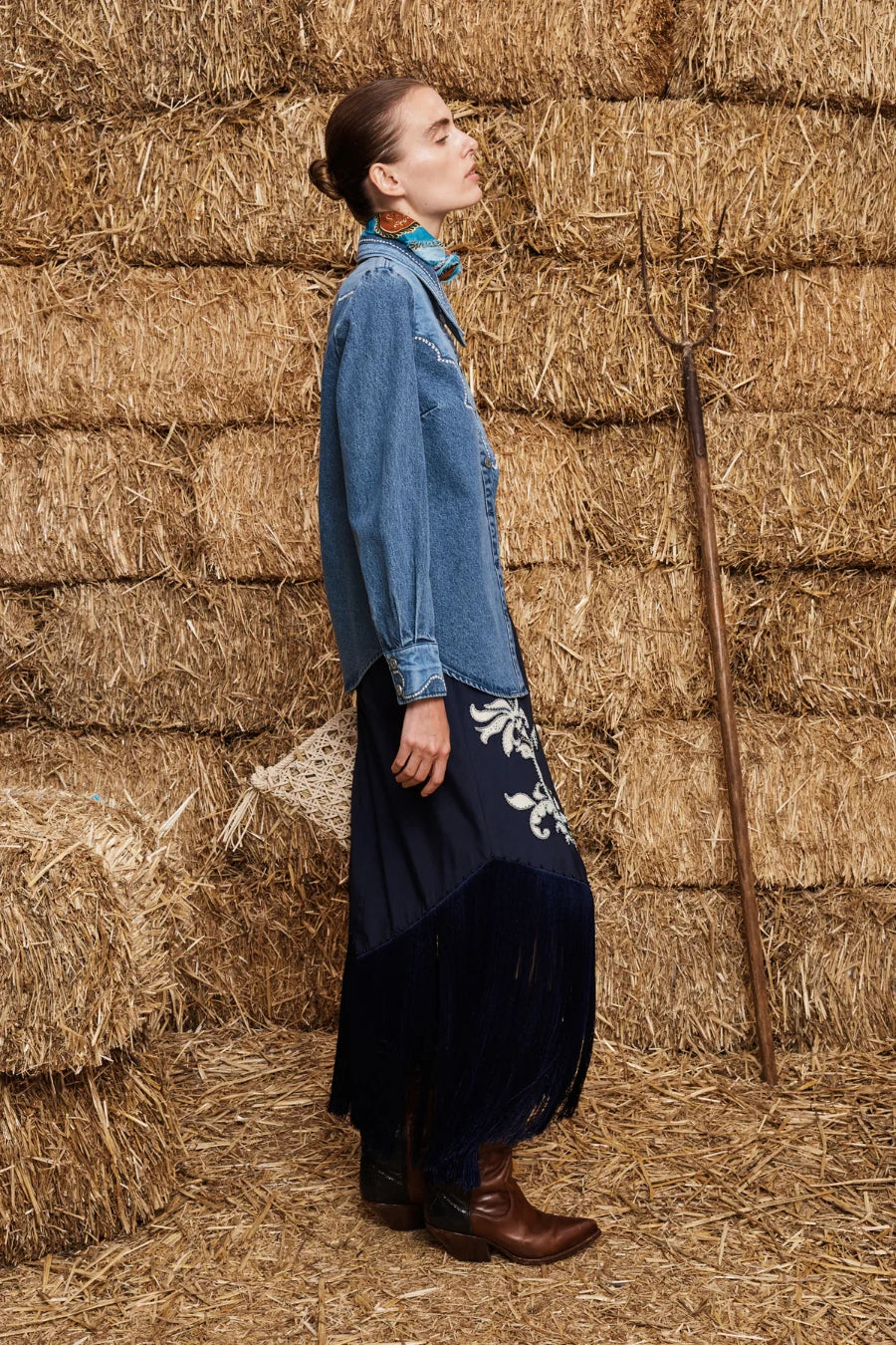 A woman stands in profile against a backdrop of stacked hay bales, wearing a long-sleeve blue denim shirt dress with a dark navy skirt featuring white floral embroidery, paired with brown leather boots.