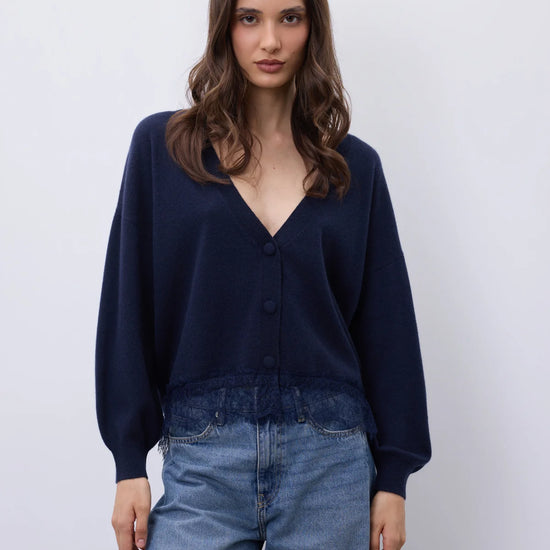 A woman modeling a dark navy blue button-up cardigan sweater with a V-neck and long sleeves, paired with light blue jeans, against a plain light gray background.