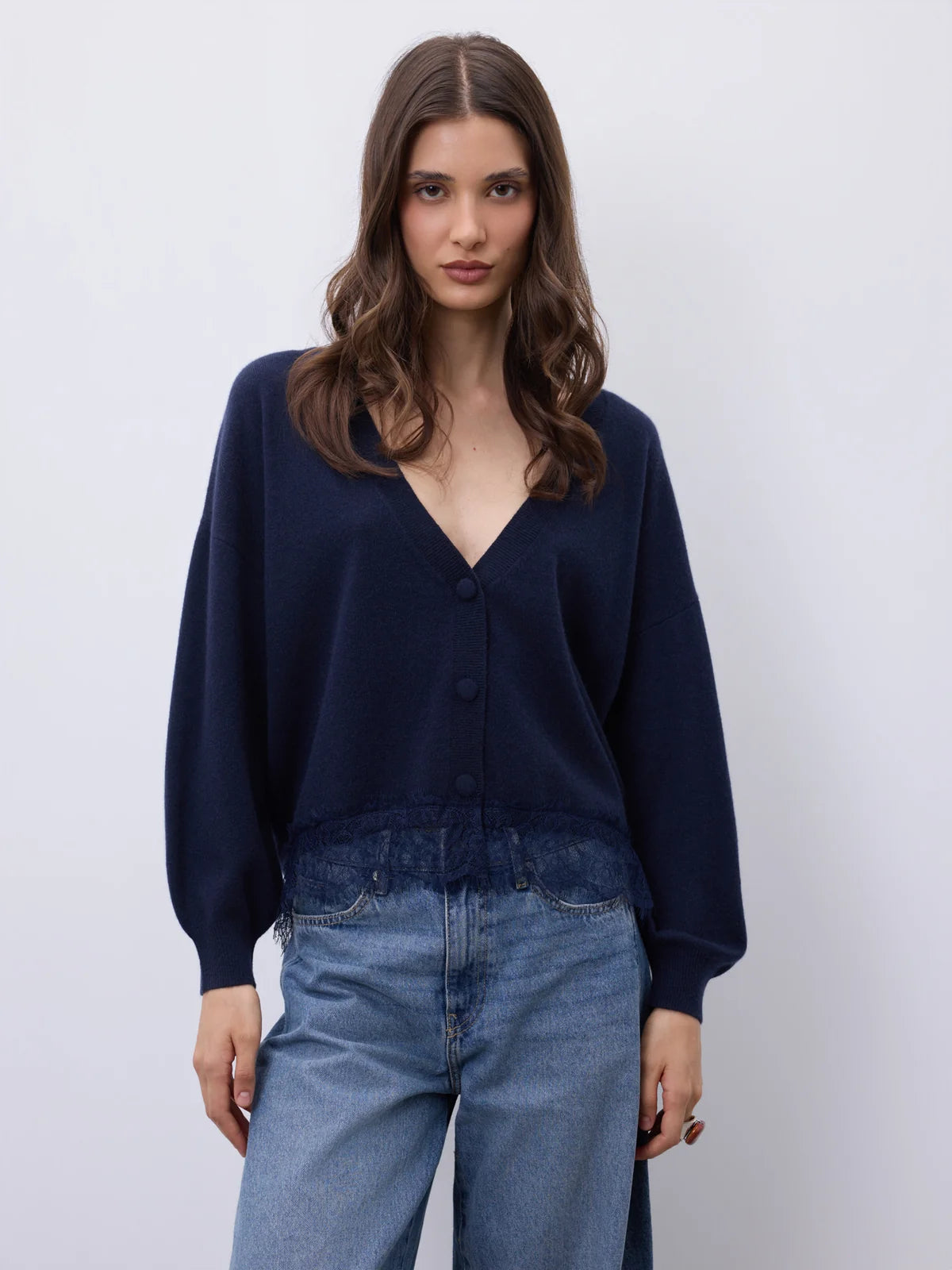 A woman modeling a dark navy blue button-up cardigan sweater with a V-neck and long sleeves, paired with light blue jeans, against a plain light gray background.