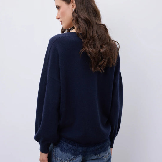 A woman with long brown hair is shown from the back wearing a dark navy blue knit sweater and blue jeans against a plain light gray background.