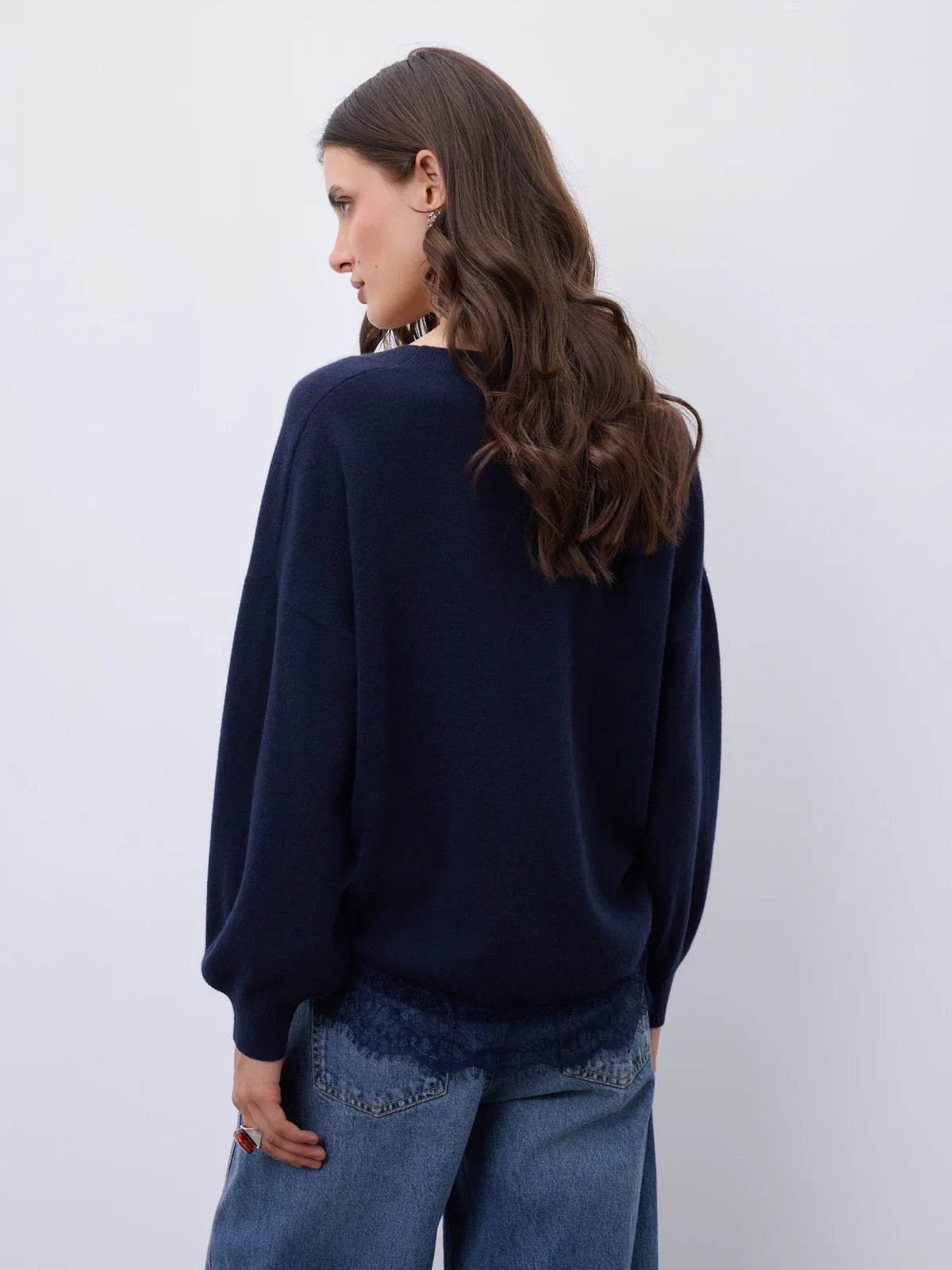 A woman with long brown hair is shown from the back wearing a dark navy blue knit sweater and blue jeans against a plain light gray background.