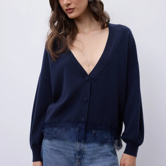A woman wearing a dark navy blue knit cardigan with a deep V-neck and button-front closure, paired with light blue jeans, photographed against a plain light gray studio background.