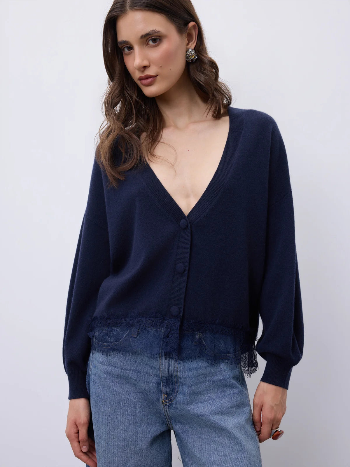 A woman wearing a dark navy blue knit cardigan with a deep V-neck and button-front closure, paired with light blue jeans, photographed against a plain light gray studio background.