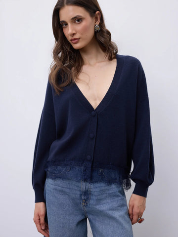 A woman wearing a dark navy blue knit cardigan with a deep V-neck and button-front closure, paired with light blue jeans, photographed against a plain light gray studio background.