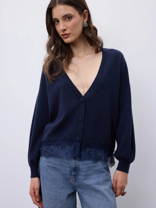 A woman wearing a dark navy blue knit cardigan with a deep V-neck and button-front closure, paired with light blue jeans, photographed against a plain light gray studio background.