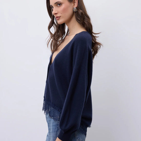 A woman wearing a navy blue knit cardigan with fringe detailing and long sleeves, paired with blue jeans, photographed against a plain light gray background.