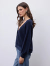 A woman wearing a navy blue knit cardigan with fringe detailing and long sleeves, paired with blue jeans, photographed against a plain light gray background.