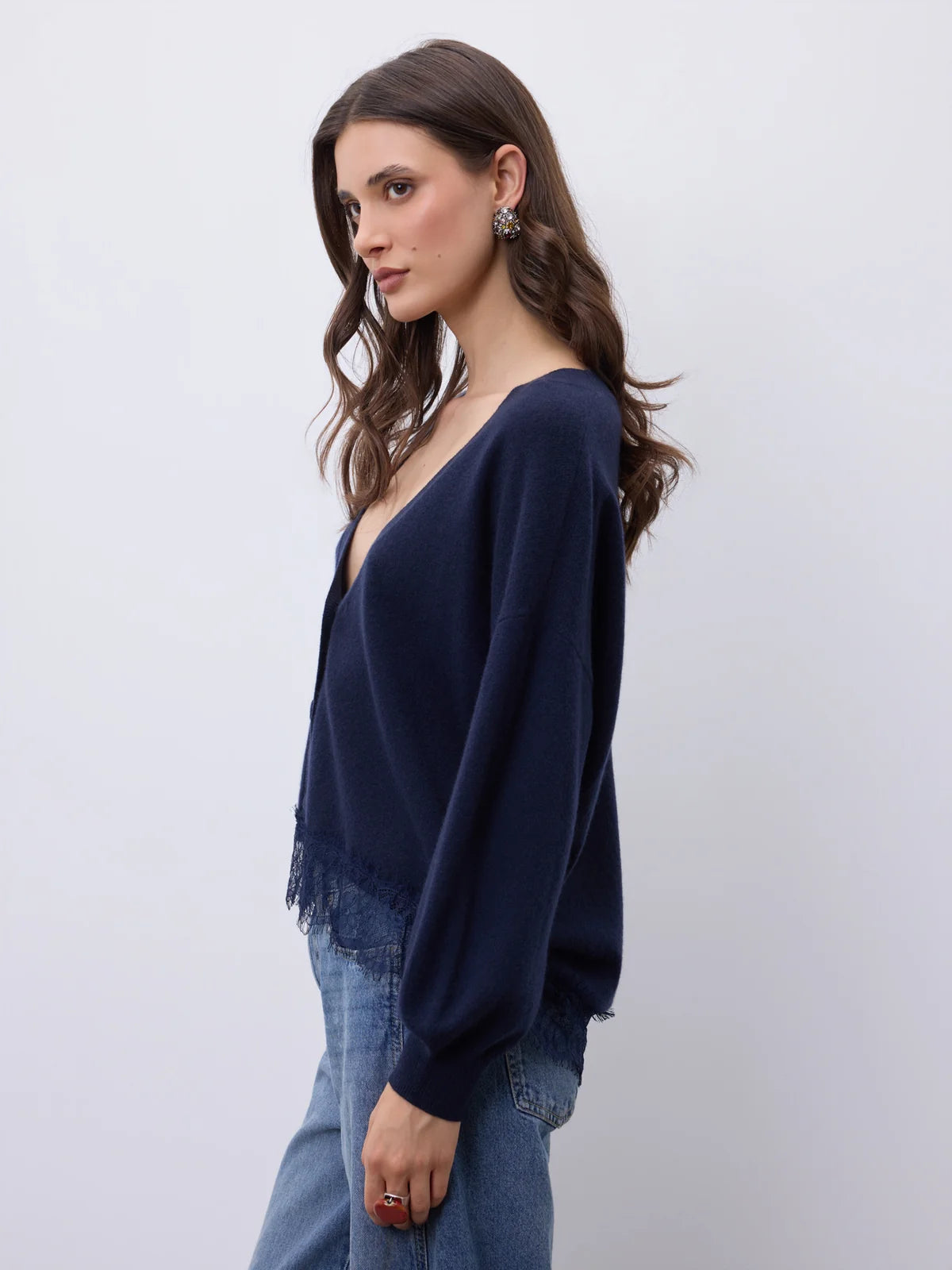 A woman wearing a navy blue knit cardigan with fringe detailing and long sleeves, paired with blue jeans, photographed against a plain light gray background.