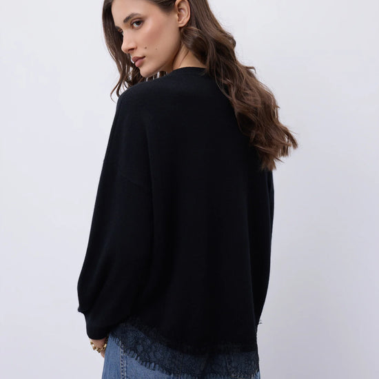 A woman wearing a black knit sweater and blue jeans, photographed from the back against a plain light gray studio background.