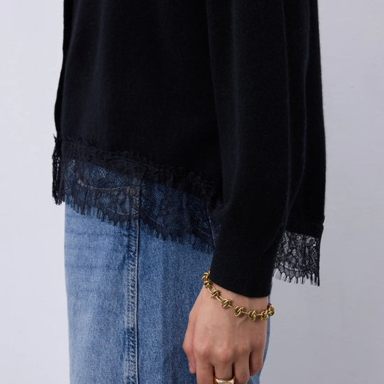 A person wearing a black long-sleeve top with lace trim at the hem and cuffs, paired with a blue denim skirt. The person is also wearing a gold chain bracelet and a gold ring, shown against a plain light gray background.