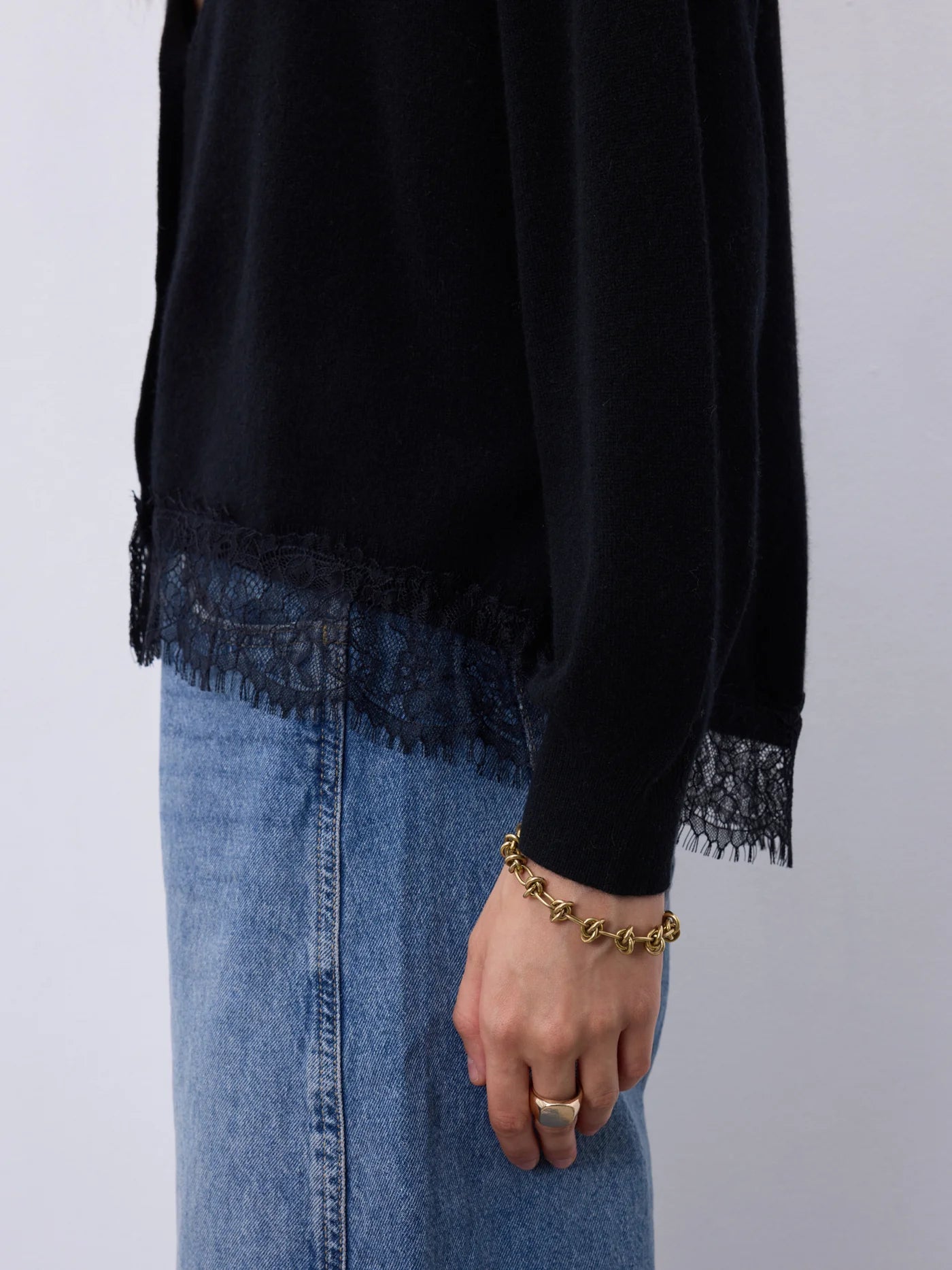A person wearing a black long-sleeve top with lace trim at the hem and cuffs, paired with a blue denim skirt. The person is also wearing a gold chain bracelet and a gold ring, shown against a plain light gray background.