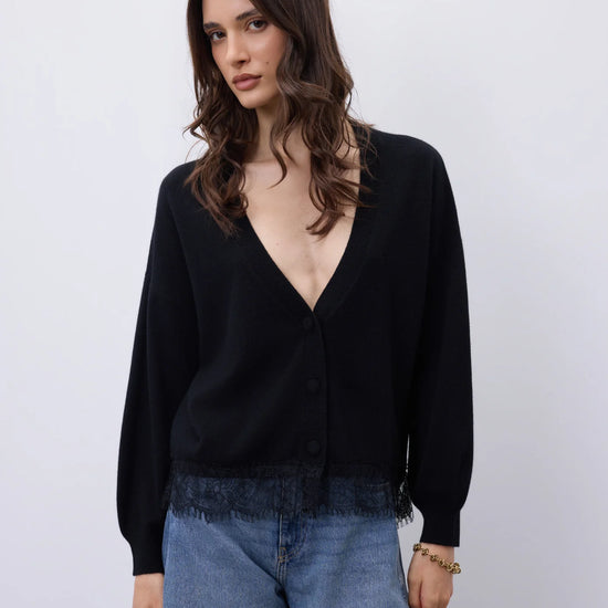 A woman wearing a black long-sleeve cardigan with a deep V-neck and button front, paired with light blue jeans, against a plain light gray studio background.