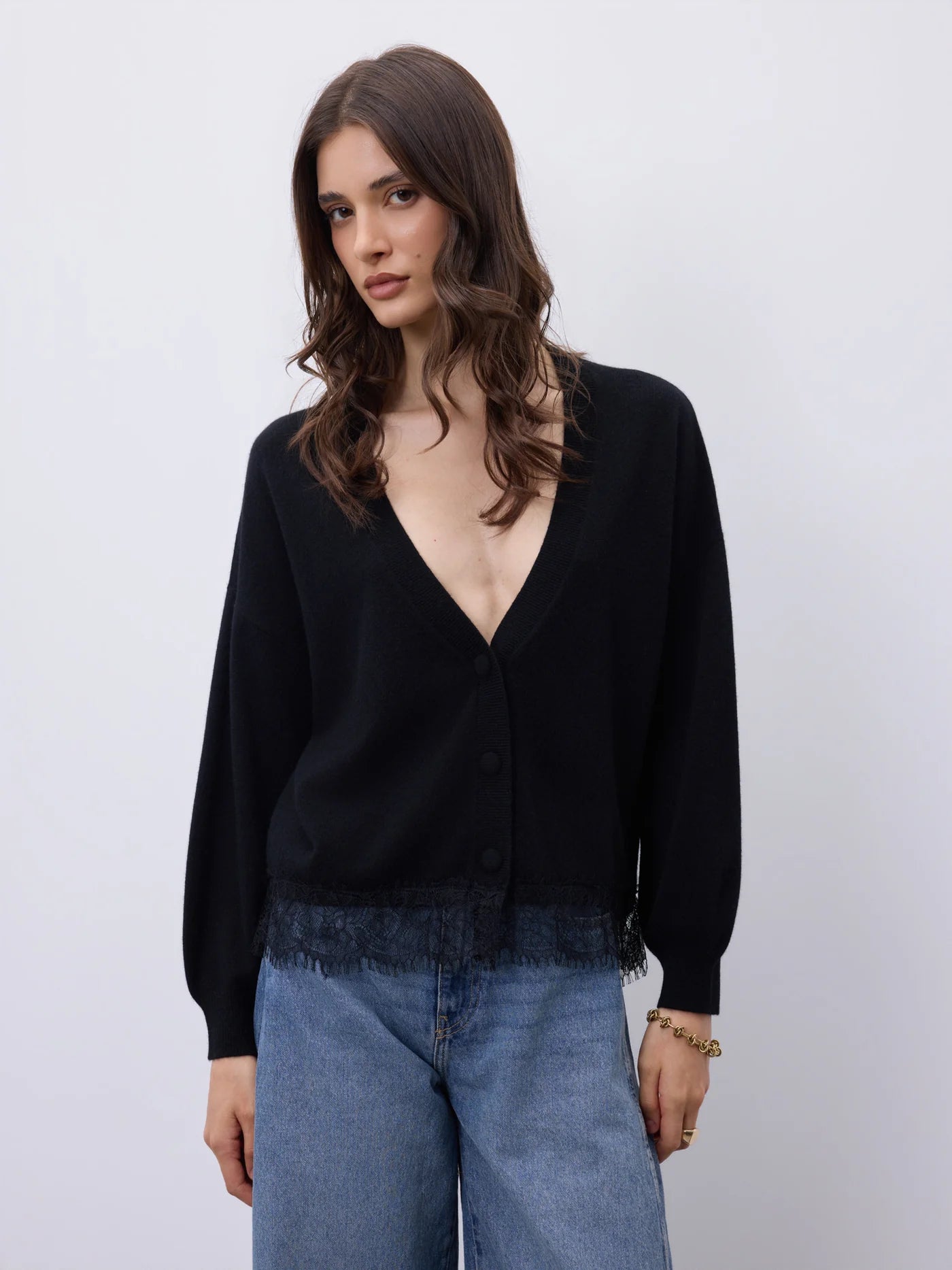 A woman wearing a black long-sleeve cardigan with a deep V-neck and button front, paired with light blue jeans, against a plain light gray studio background.