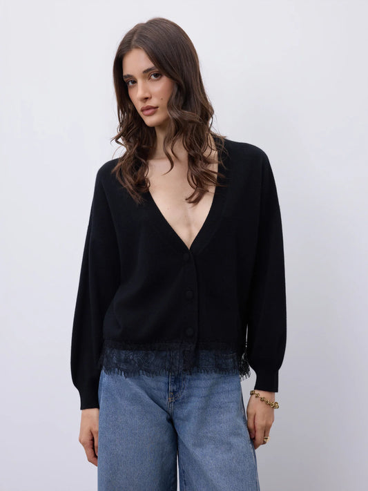 A woman wearing a black long-sleeve cardigan with a deep V-neck and button front, paired with light blue jeans, against a plain light gray studio background.