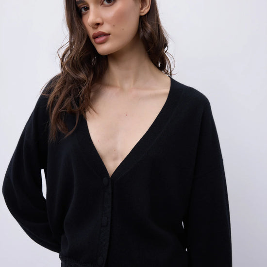 A woman wearing a black long-sleeve V-neck cardigan with lace trim at the hem, paired with blue jeans, against a plain light gray background.
