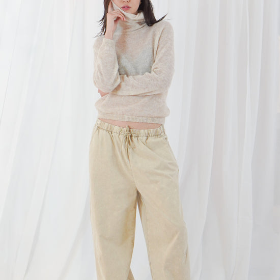 A woman stands barefoot against a white draped fabric backdrop, wearing a light beige turtleneck sweater and matching beige wide-leg pants.