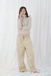 A woman stands barefoot against a white draped fabric backdrop, wearing a light beige turtleneck sweater and matching beige wide-leg pants.
