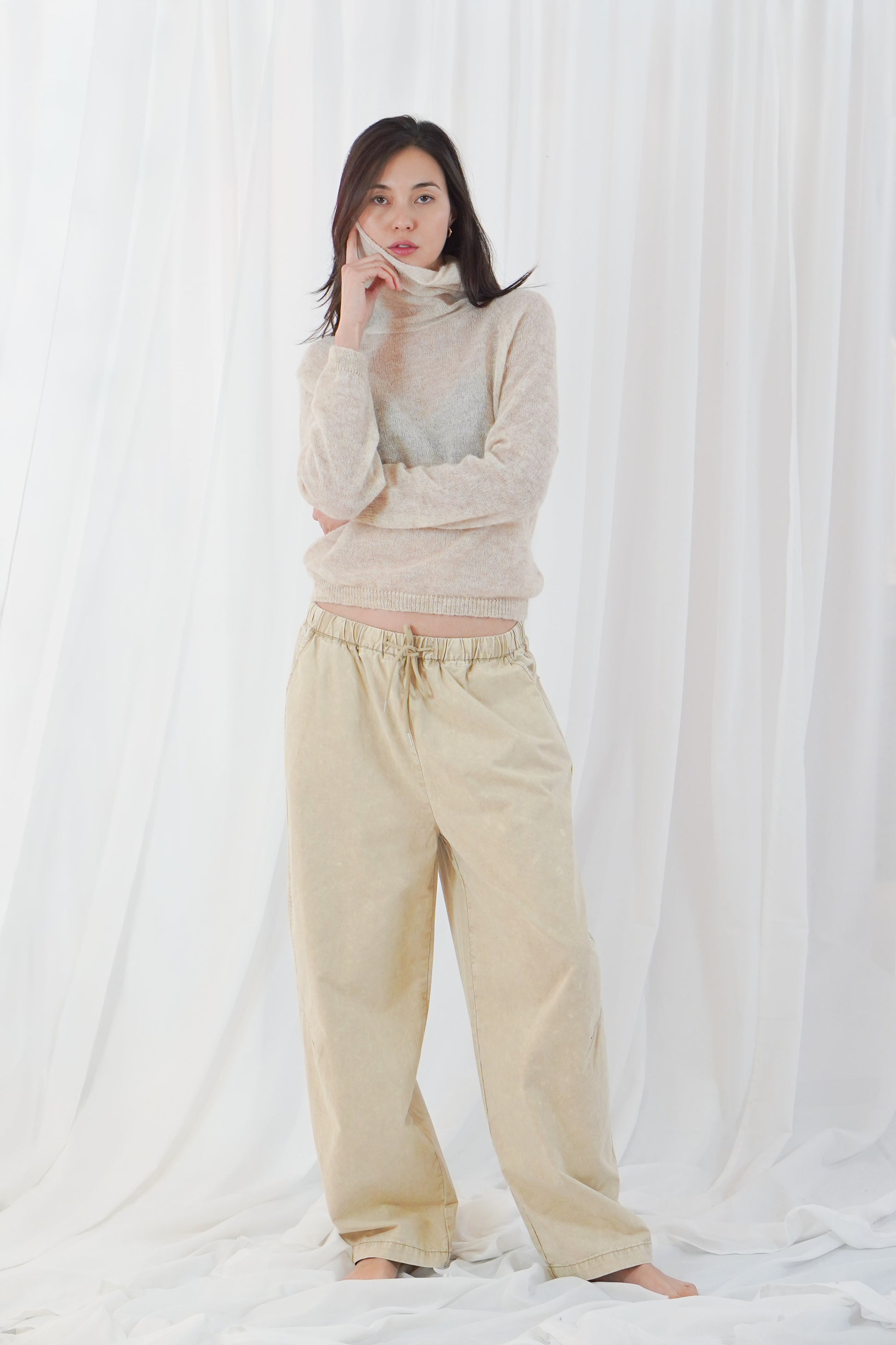 A woman stands barefoot against a white draped fabric backdrop, wearing a light beige turtleneck sweater and matching beige wide-leg pants.