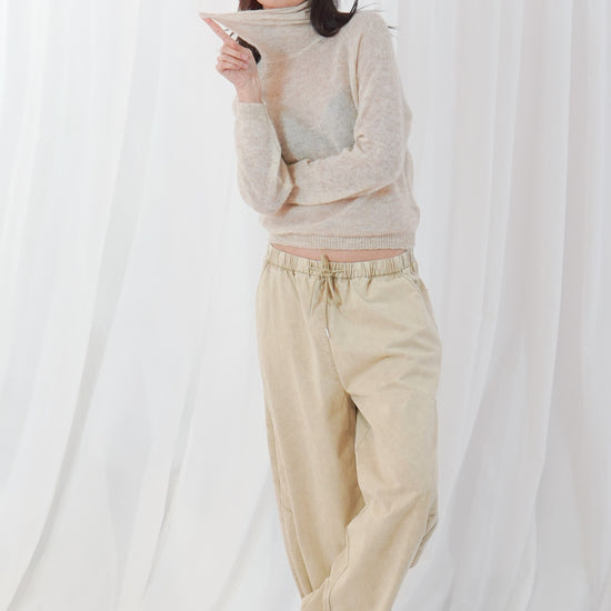 A woman stands barefoot in front of a white draped fabric backdrop, wearing a loose beige turtleneck sweater and matching beige drawstring pants.