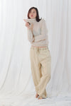A woman stands barefoot in front of a white draped fabric backdrop, wearing a loose beige turtleneck sweater and matching beige drawstring pants.