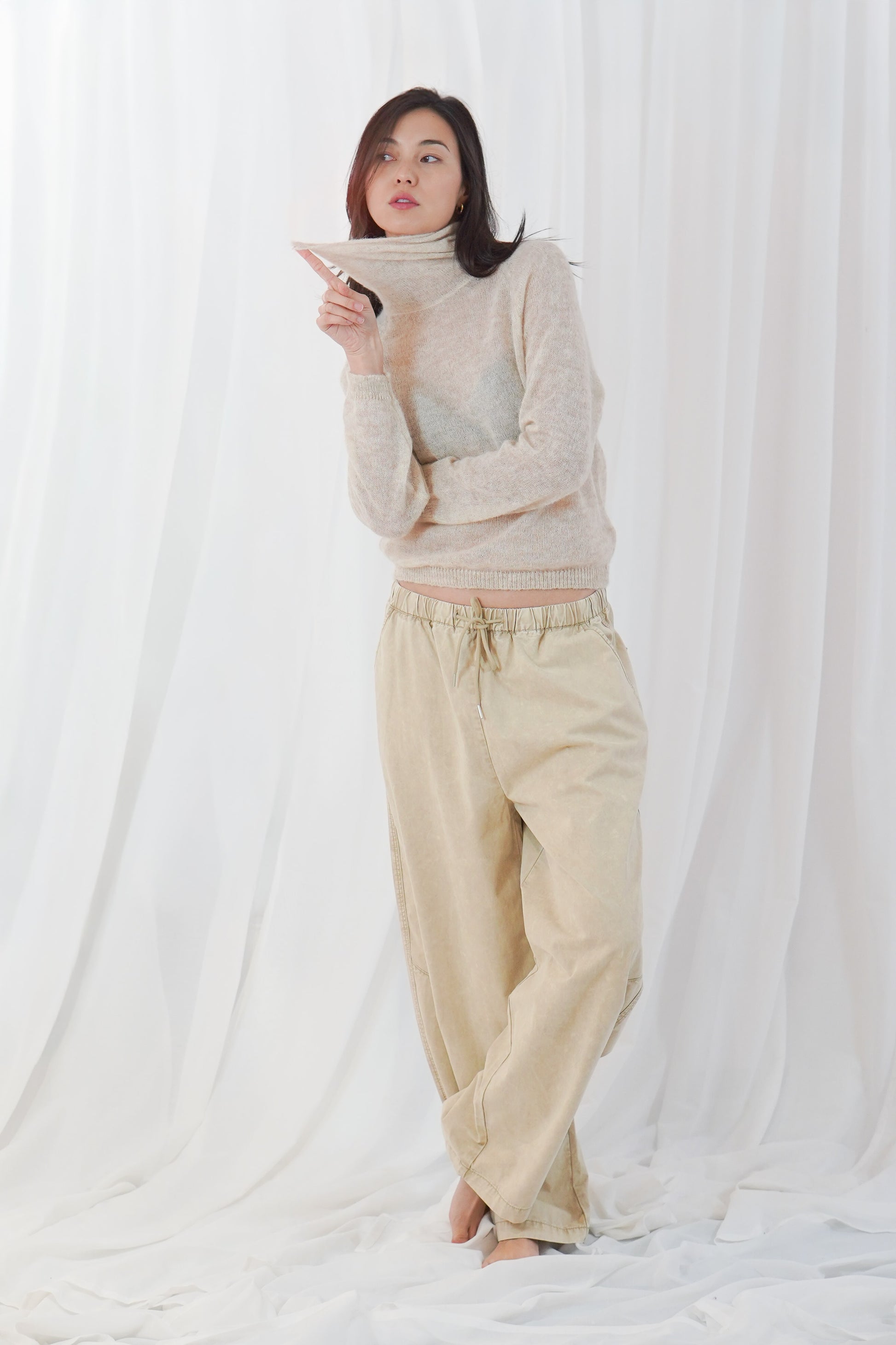 A woman stands barefoot in front of a white draped fabric backdrop, wearing a loose beige turtleneck sweater and matching beige drawstring pants.
