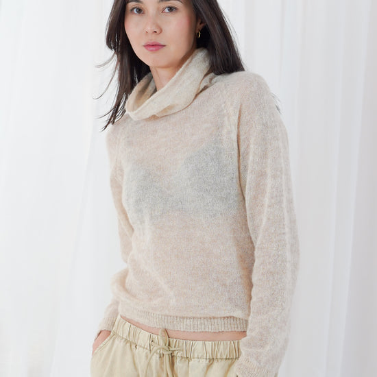 A woman wearing a fuzzy, light beige cowl-neck sweater and matching beige drawstring trousers, standing against a white curtain backdrop.